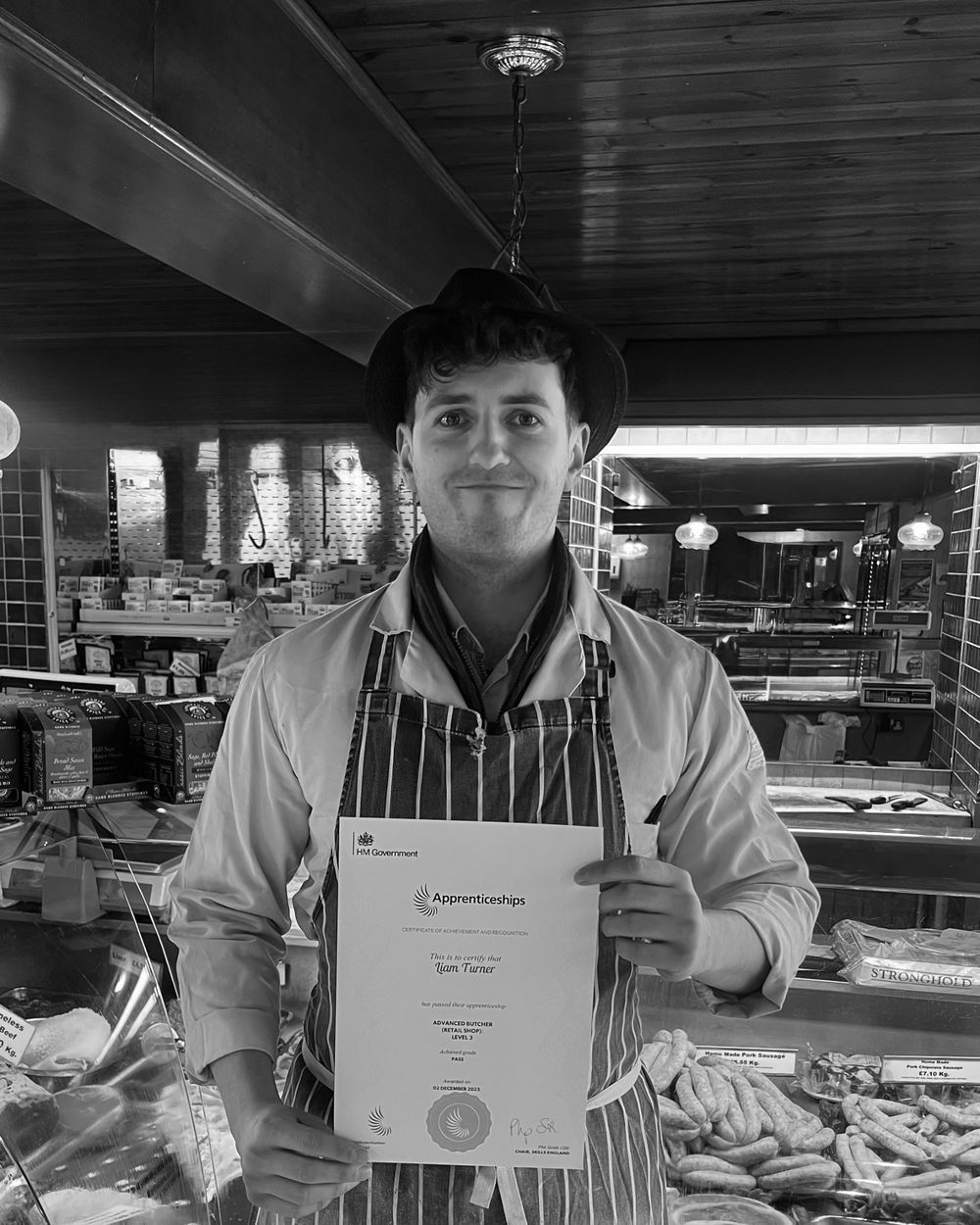 Black and white portrait of Liam Turner in his butcher’s apron and hat, holding his Advanced Butcher apprenticeship certificate